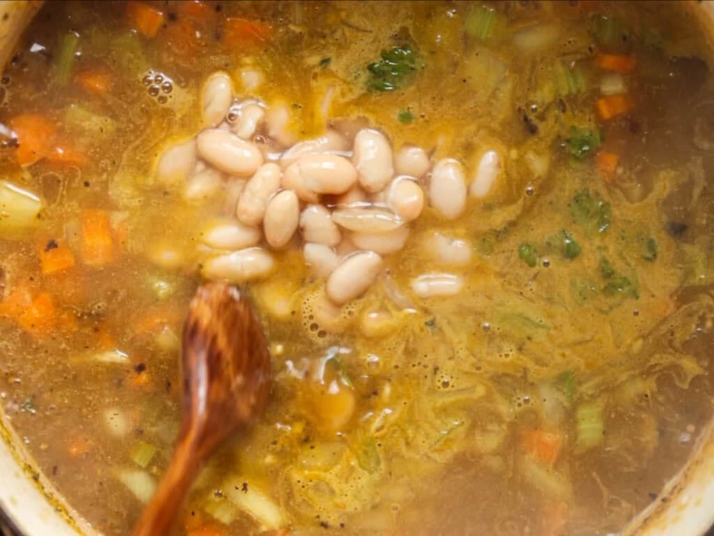 A wooden spoon stirs a pot of soup with white beans, diced carrots, celery, and herbs in a golden broth.